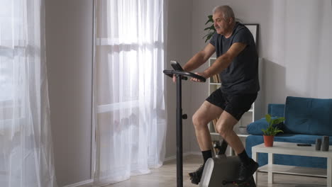 sporty-middle-aged-man-is-training-with-stationary-bicycle-at-home-medium-shot-in-living-room-healthy-lifestyle-in-old-age